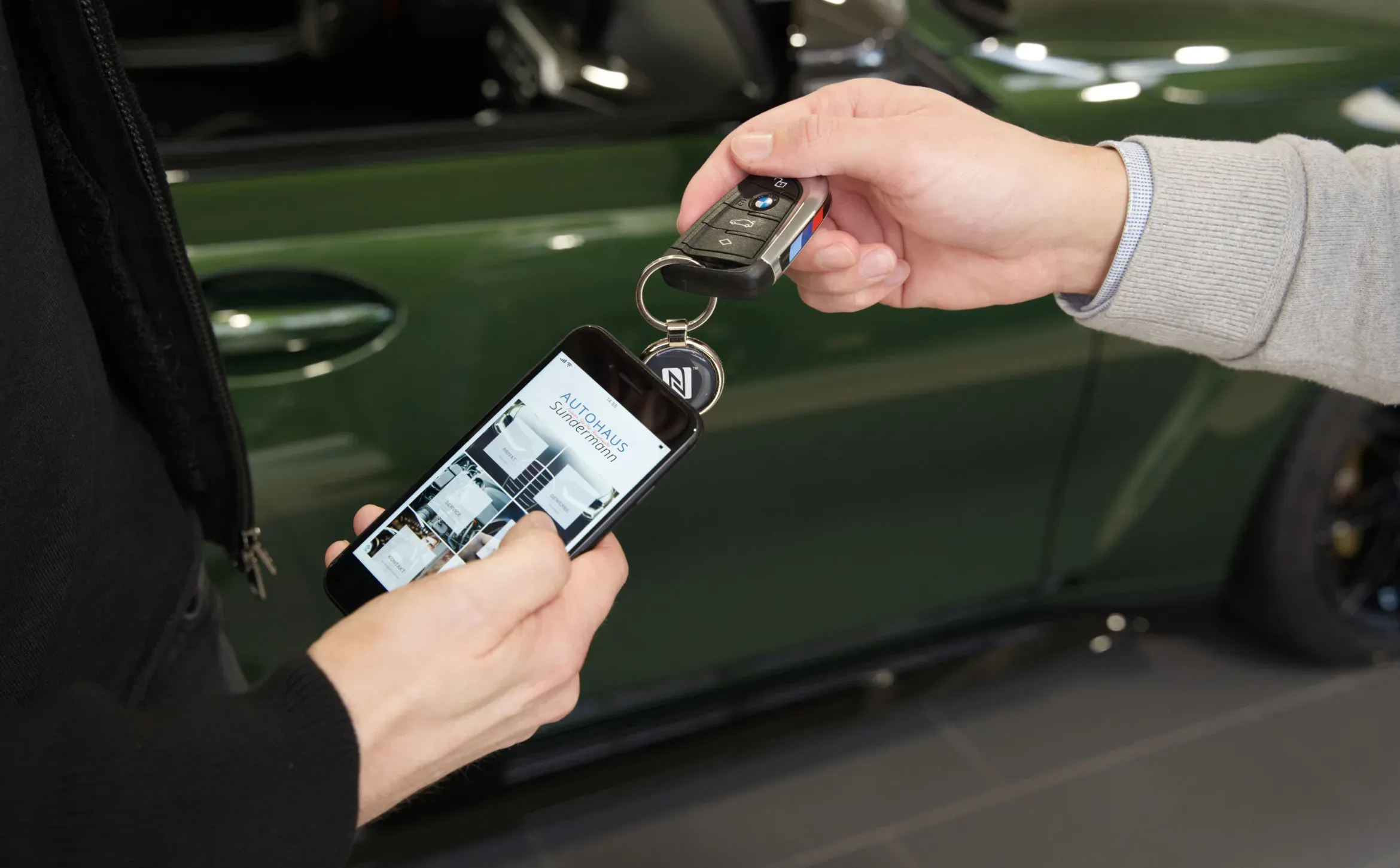 Auf dem Bild sieht man folgendes: Ein NFC-Anhänger, der an einem Autoschlüssel befestigt ist, wird mit einem Smartphone gescannt. Auf dem Smartphone erscheinen dann Informationen über das Autohaus.